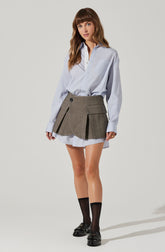 A woman is wearing an oversized, light blue, long-sleeved button-up shirt with subtle vertical stripes. The shirt has a collar and a chest pocket, and it is tucked into a high-waisted, khaki pleated mini skirt that features a large flap pocket on the right side, a zippered slit detail on the left, and a single button on the waistband. She is also wearing black socks that reach mid-calf and black loafers with metallic buckle accents.